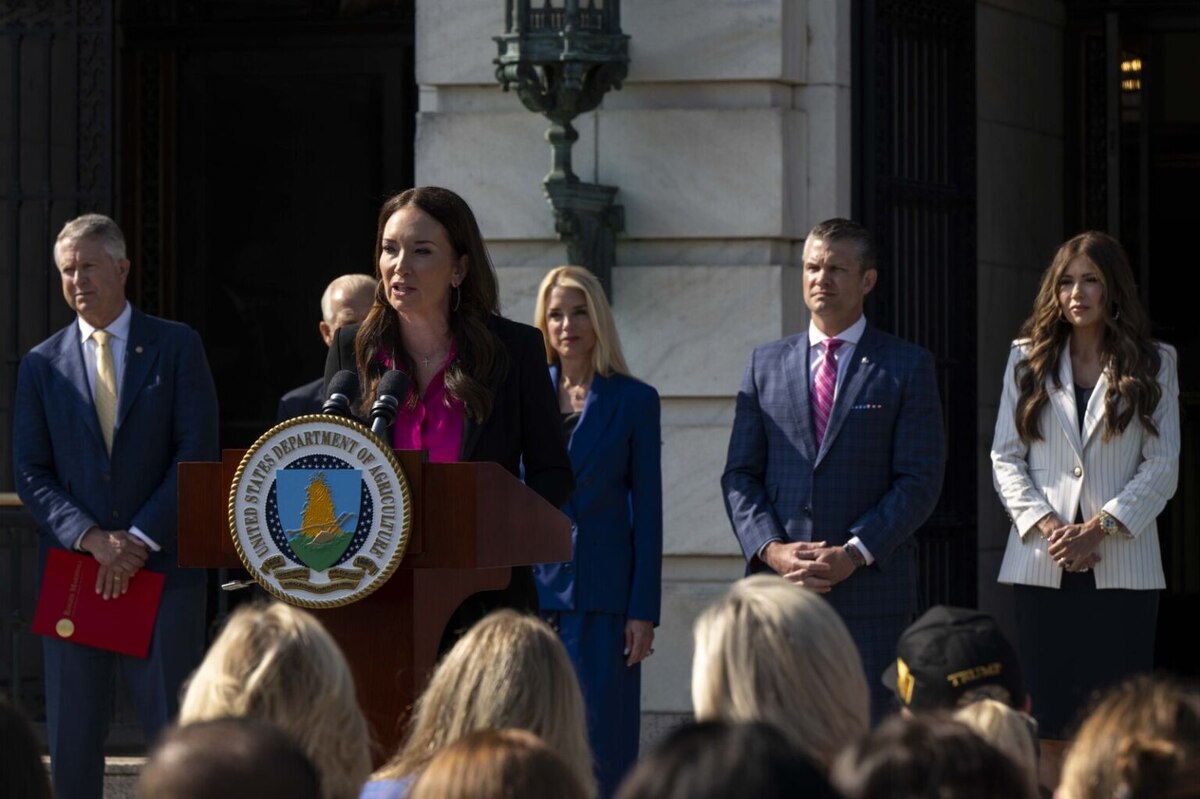A woman in business attire speaks at a podium, with a group of men and women in similar attire standing behind her.