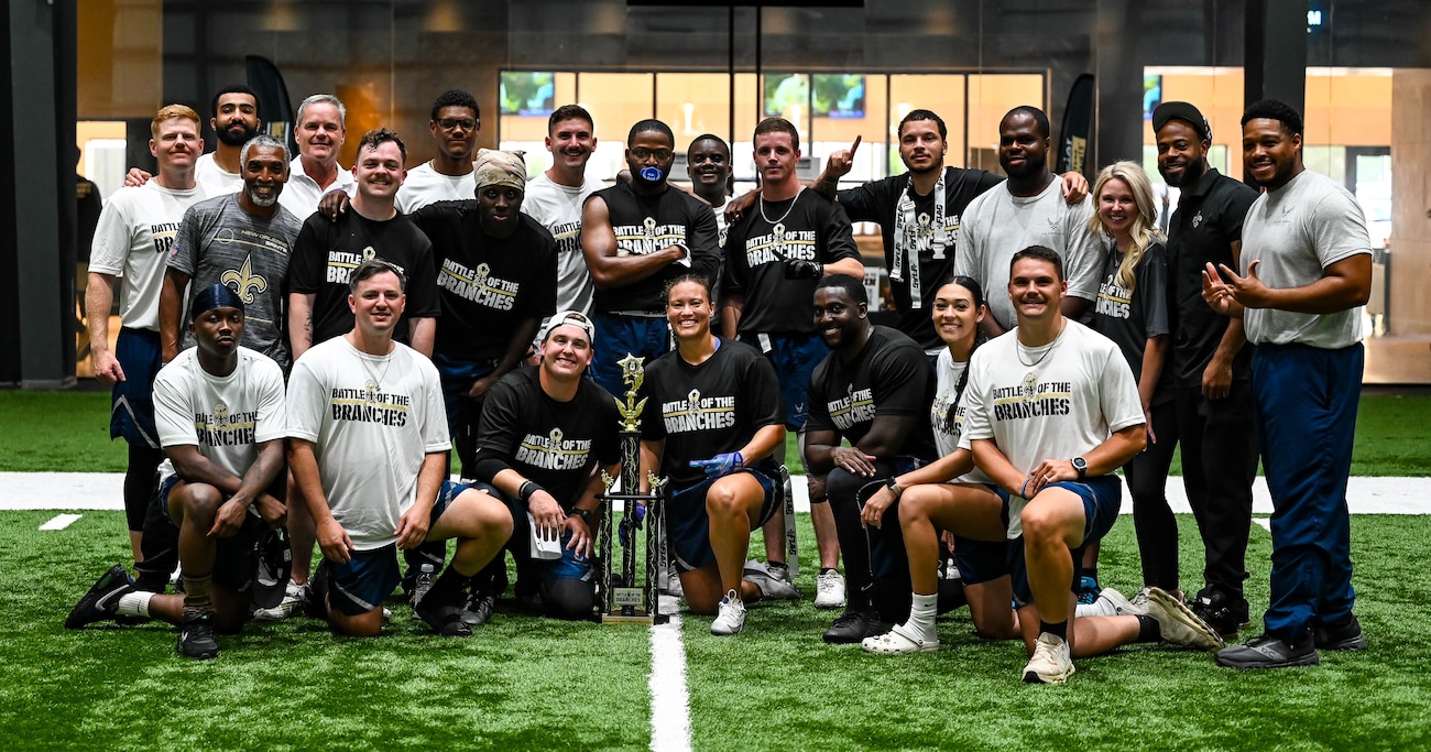 A group of people in athletic gear smiling for a photo with a black and gold trophy on a green turf indoor football field.