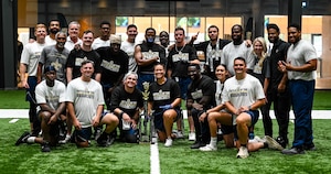 A group of people in athletic gear smiling for a photo with a black and gold trophy on a green turf indoor football field.