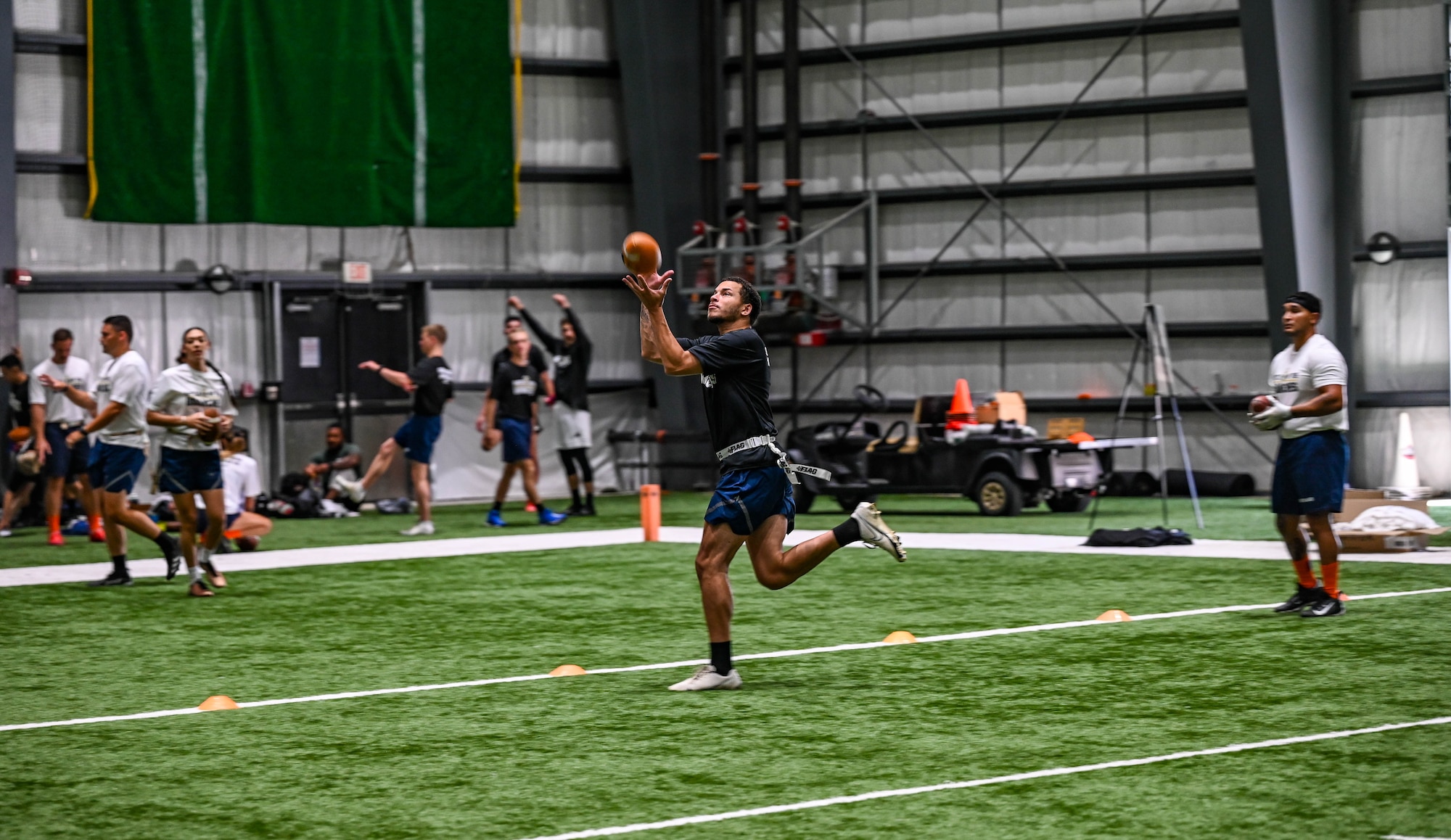 A man in blue shorts and a black shirt catches a football on an indoor turf field.
