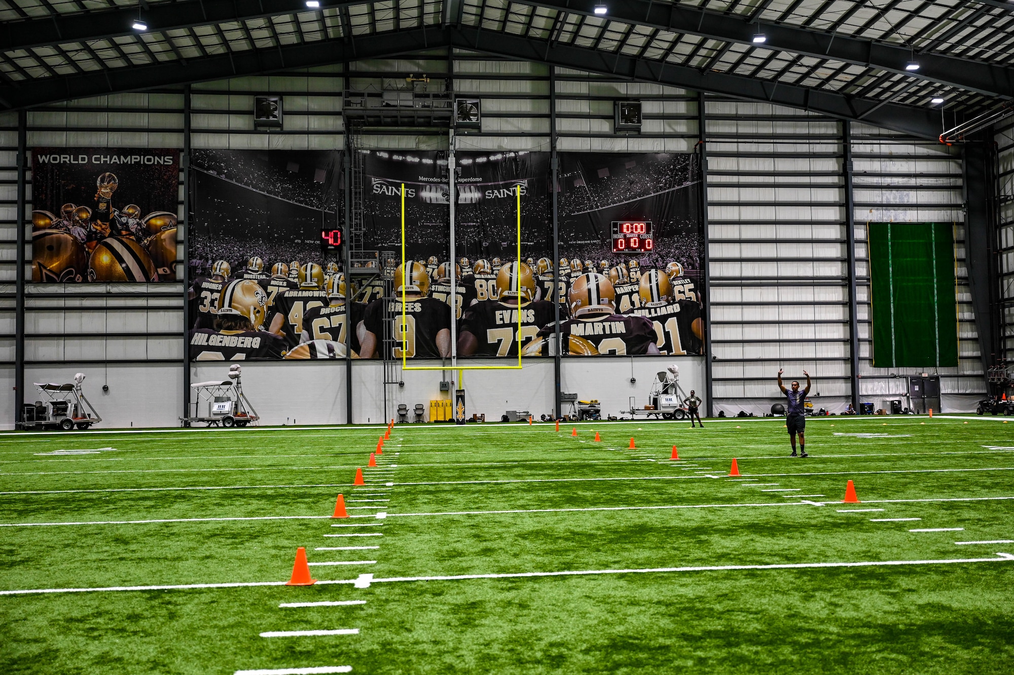 A wide shot of the New Orleans Saints indoor practice facility with a green turf football field, a yellow goalpost, and orange cones lining the field.