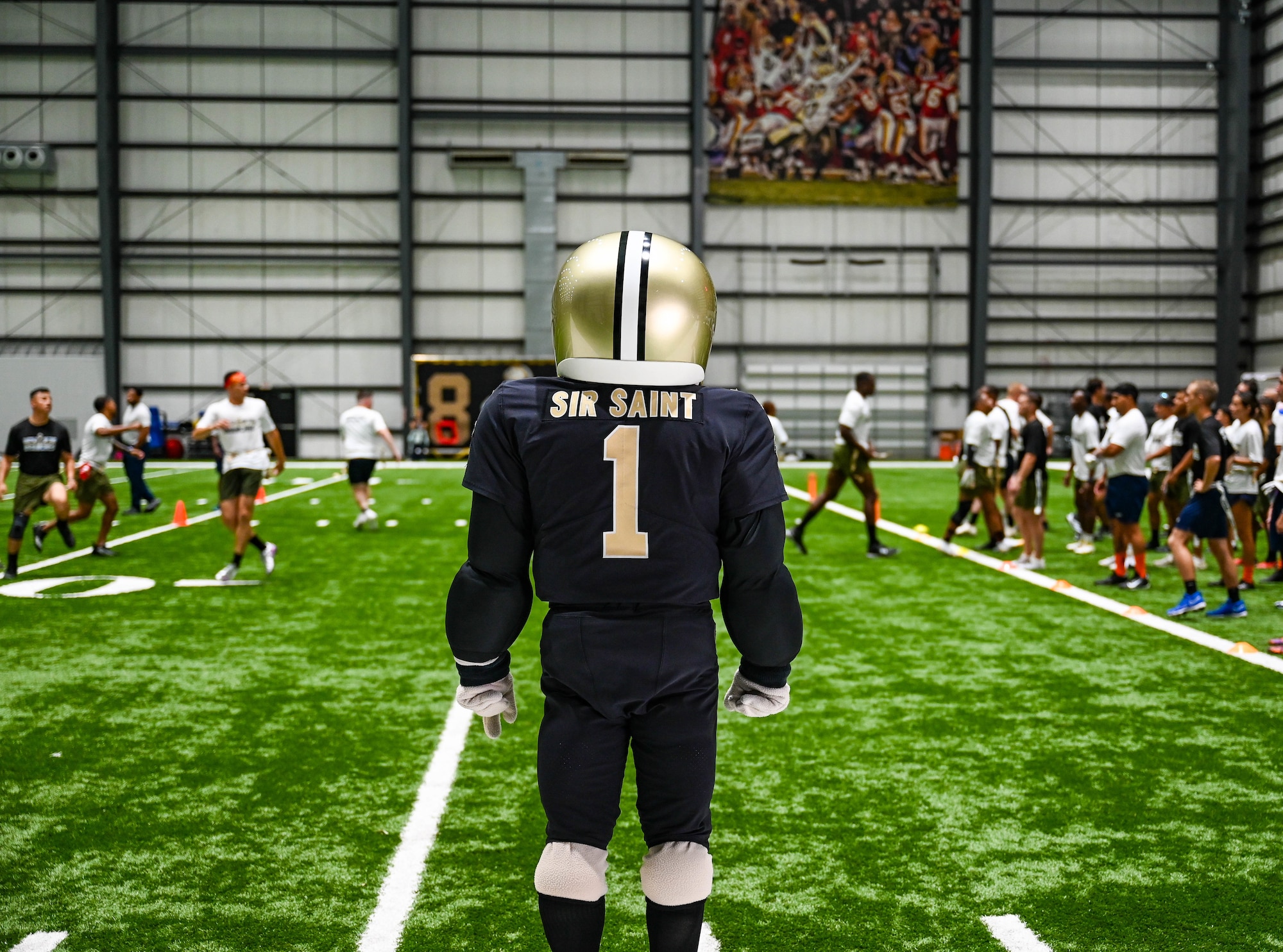 A mascot of a New Orleans Saints player stands on a green turf indoor field.