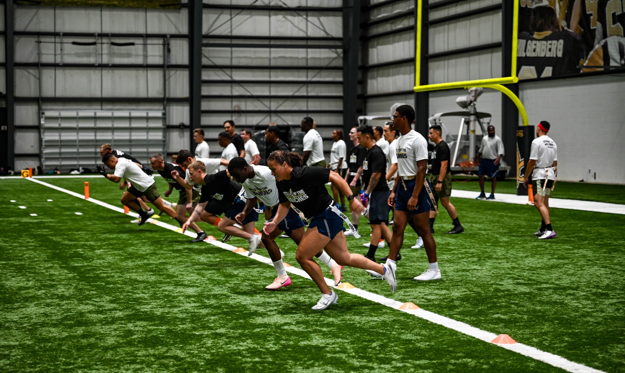 A group of military personnel in athletic gear run across a green turf indoor football field.