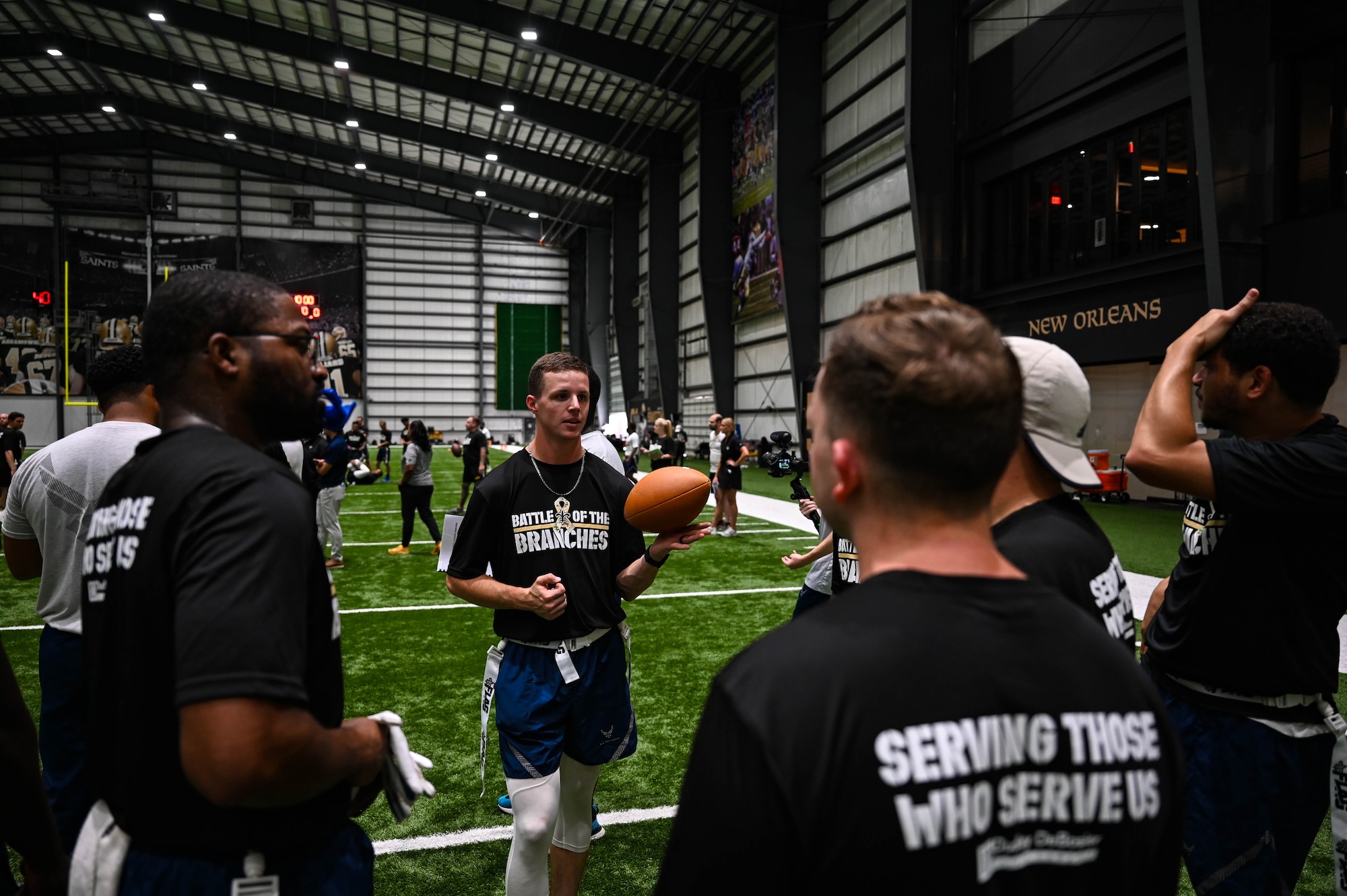 A man in athletic gear holding a football while talking to his team on a green turf indoor football field.