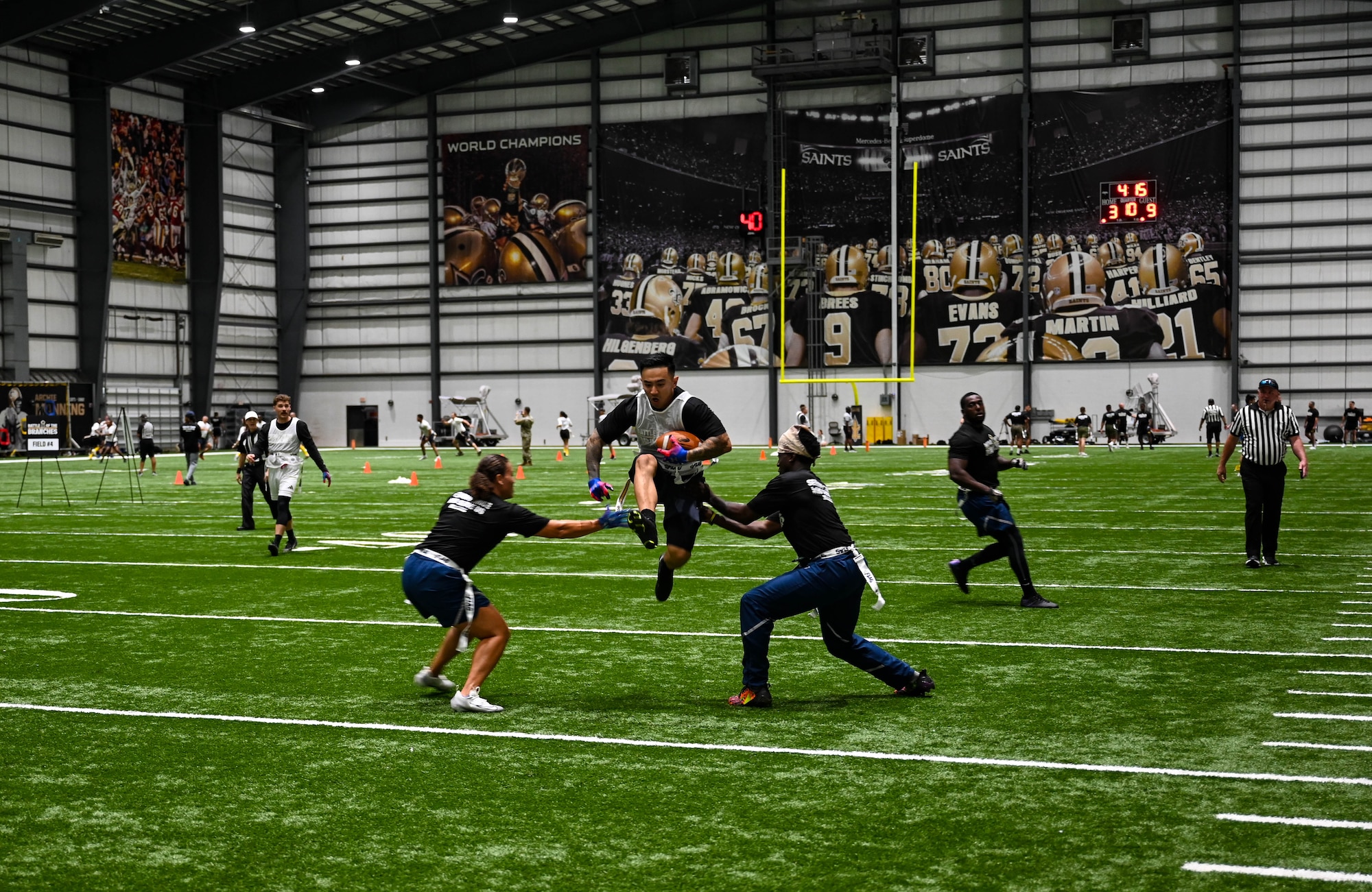 A man in athletic gear and a football tucked into his arm jumping over two other people in athletic gear on a green turf indoor football field.