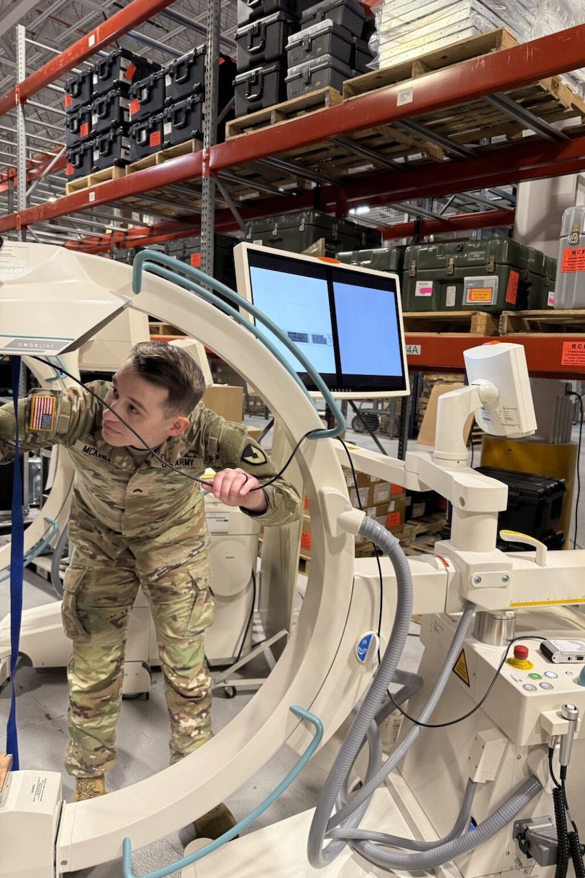 A C-shaped X-ray machine is in a warehouse being inspected by a soldier in a camouflage uniform.