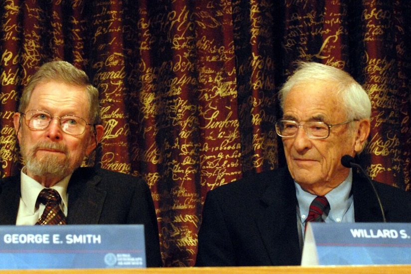 Two men in suits are seated with nameplates and microphones in front of them.