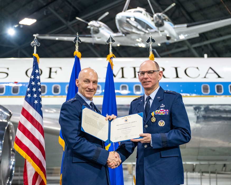 Two Air Force officers pose for a photo in service dress uniforms in front of U.S. and military flags