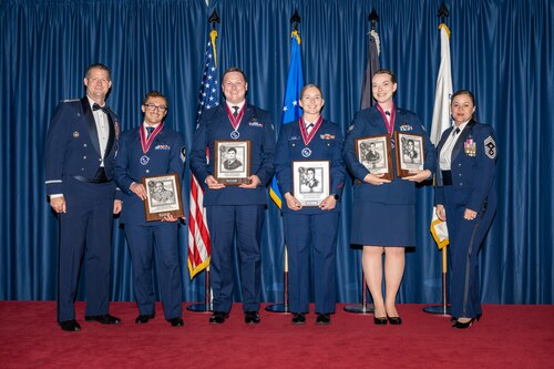 Six Air Force service members pose for a photo wearing service dress uniforms and holding award plaques