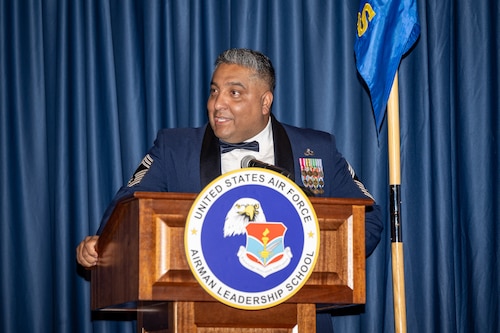 A man in Air Force service dress uniform stands behind a podium with an emblem for the Airman Leadership School