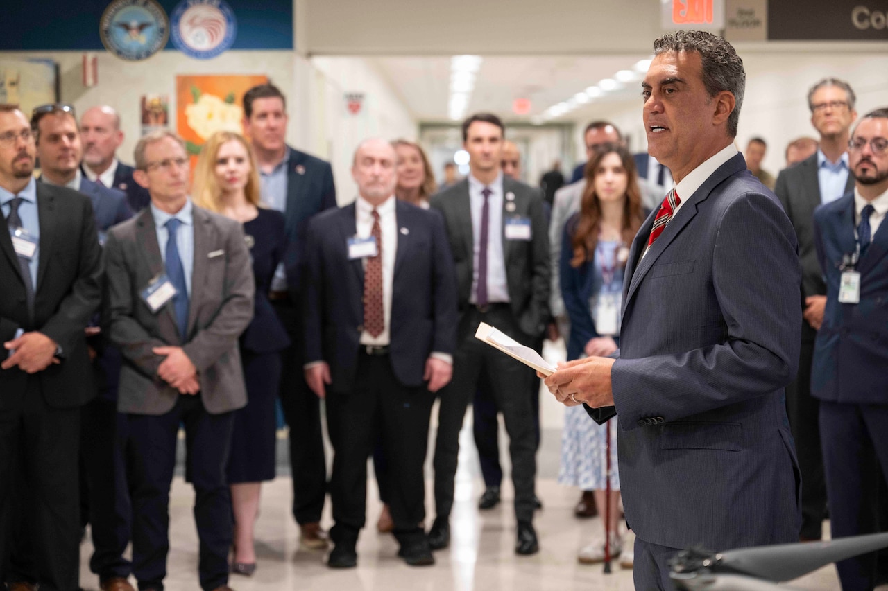 A man in a suit speaks to a crowd of people in business attire inside a building.