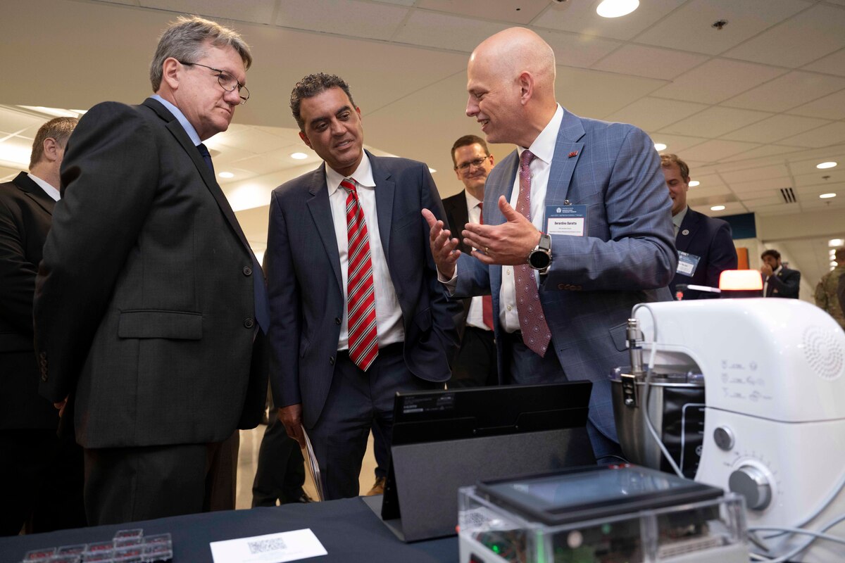 Three men in suits speak while looking at advanced manufacturing technology.