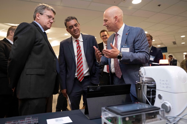 Three men in suits speak while looking at advanced manufacturing technology.
