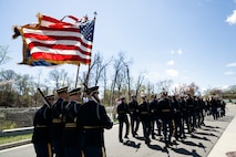 Army Soldiers dressed in ceremonial uniforms are marching (away from the camera) in formation down a street, with the back color guard formation of four Soldiers carrying the US flag, the Army flag and two with rifles on each end.