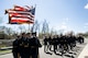 Army Soldiers dressed in ceremonial uniforms are marching (away from the camera) in formation down a street, with the back color guard formation of four Soldiers carrying the US flag, the Army flag and two with rifles on each end.