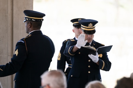 An Army Soldier wearing a dark ceremonial uniform with white gloves is raising his right hand, palm facing out, while reading from a black book that is in his left hand during a funeral.
