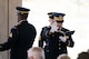 An Army Soldier wearing a dark ceremonial uniform with white gloves is raising his right hand, palm facing out, while reading from a black book that is in his left hand during a funeral.