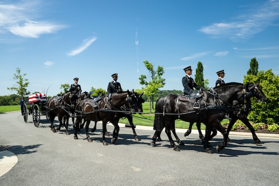Army Soldiers wearing dark ceremonial uniforms are riding on caisson horses as the caisson is carrying a flag-draped casket down a road in Arlington National Cemetery.