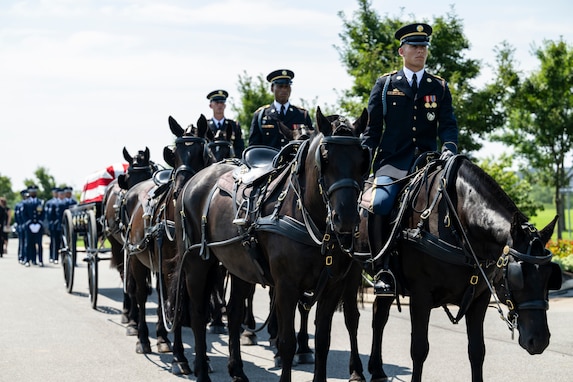 Army Soldiers wearing dark ceremonial uniforms are riding on caisson horses as the caisson is carrying a flag-draped casket down a road in Arlington National Cemetery.
