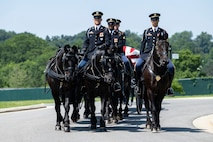 Army Soldiers are riding on caisson horses as the caisson is carrying a flag-draped casket down a road in Arlington National Cemetery.