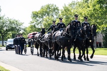 Army Soldiers are riding on caisson horses as the caisson is carrying a flag-draped casket down a road with other Soldiers marching alongside.