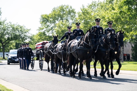 Army Soldiers are riding on caisson horses as the caisson is carrying a flag-draped casket down a road with other Soldiers marching alongside.