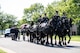 Army Soldiers are riding on caisson horses as the caisson is carrying a flag-draped casket down a road with other Soldiers marching alongside.