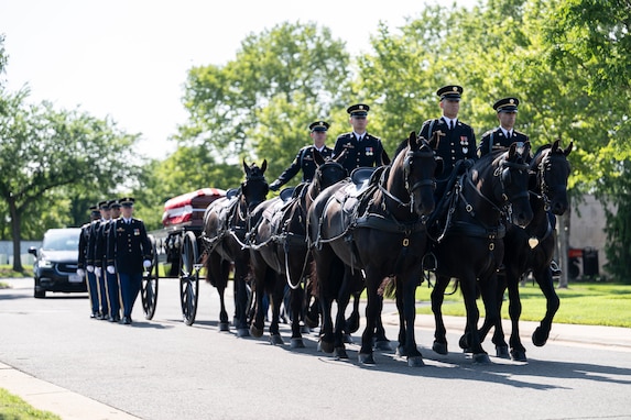 Army Soldiers are riding on caisson horses as the caisson is carrying a flag-draped casket down a road with other Soldiers marching alongside.