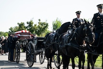 Army Soldiers from The Old Guard wearing dark ceremonial uniforms are moving a flag-draped casket from a horse-drawn caisson at Arlington National Cemetery.