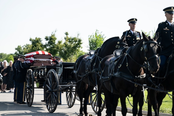 Army Soldiers from The Old Guard wearing dark ceremonial uniforms are moving a flag-draped casket from a horse-drawn caisson at Arlington National Cemetery.