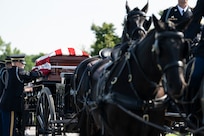 Soldiers wearing dark ceremonial uniforms are adjusting the flag that is covering a casket which is being carried on a horse-drawn caisson.