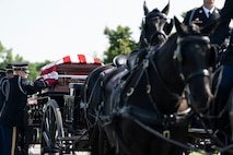 Soldiers wearing dark ceremonial uniforms are adjusting the flag that is covering a casket which is being carried on a horse-drawn caisson.