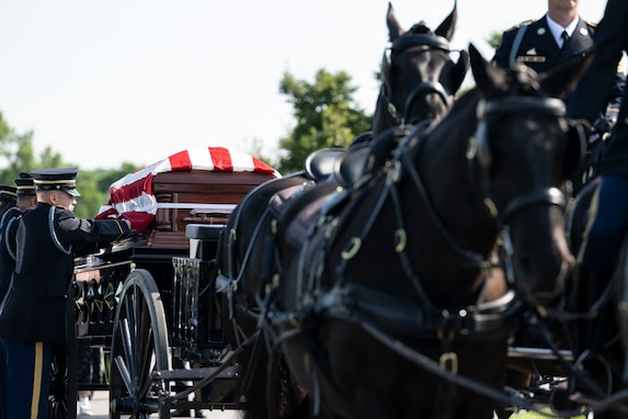 Soldiers wearing dark ceremonial uniforms are adjusting the flag that is covering a casket which is being carried on a horse-drawn caisson.
