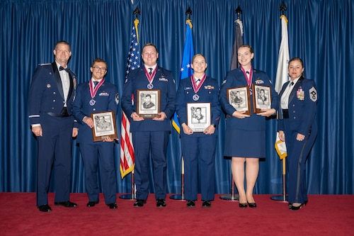 Six Air Force service members pose for a photo wearing service dress uniforms and holding award plaques
