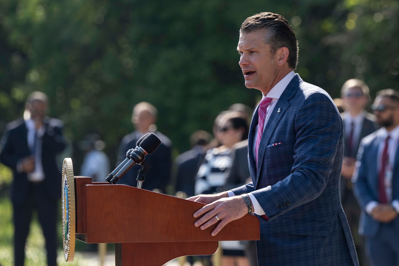 A man in  suit speaks into a microphone behind a lectern outside during the day.