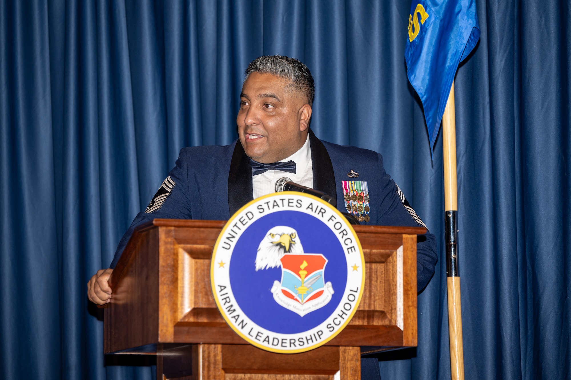 A man in Air Force service dress uniform stands behind a podium with an emblem for the Airman Leadership School