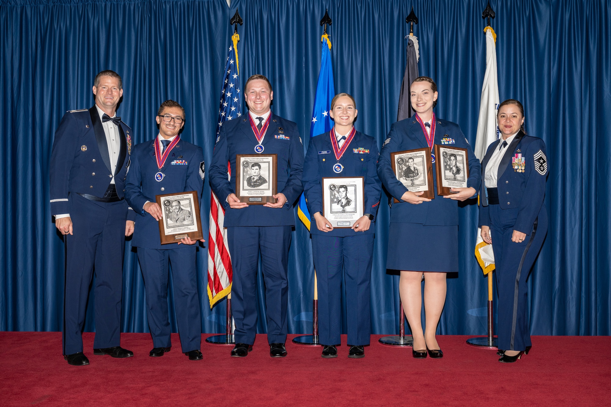 Six Air Force service members pose for a photo wearing service dress uniforms and holding award plaques