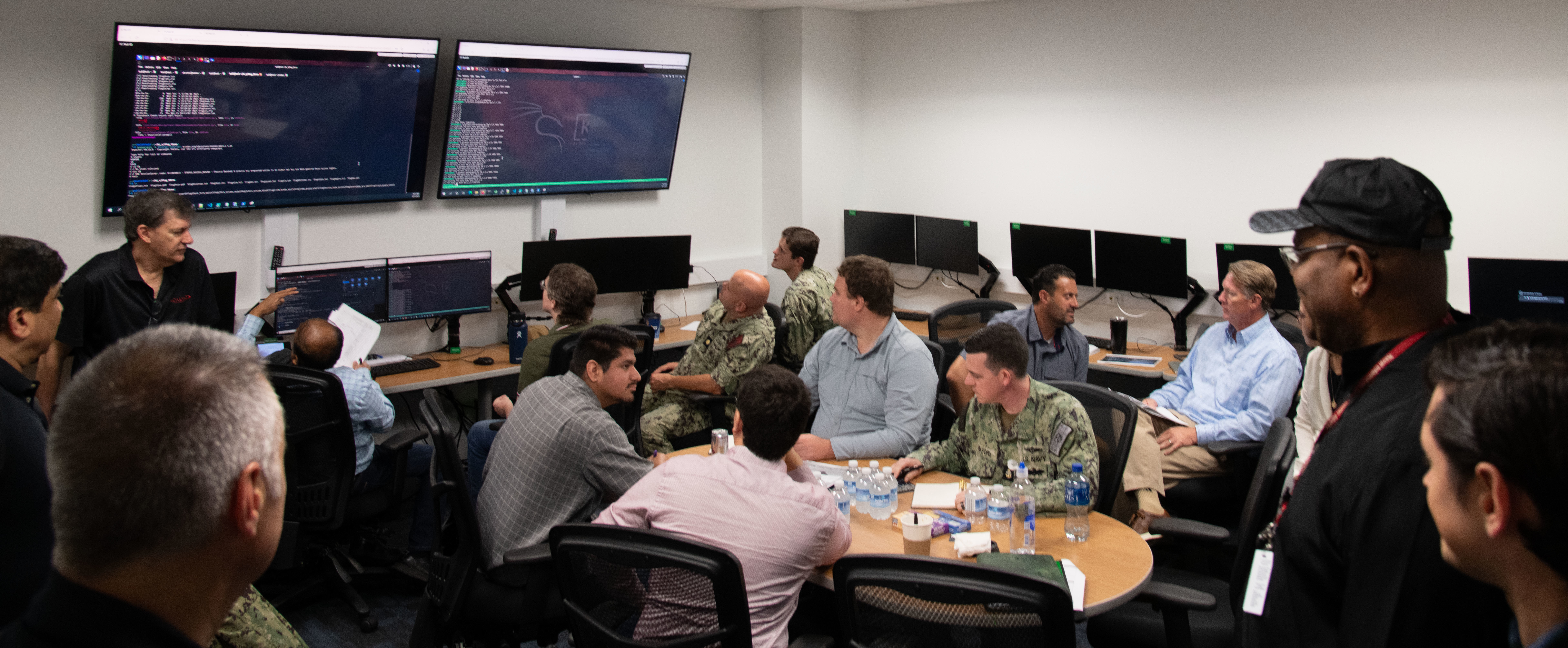 Photo of NIWC Pacific and Atlantic workforce at CyberANTX having a brief discussion.  Woman in black in front of suited men and one male navy seaman in camouflage uniform.