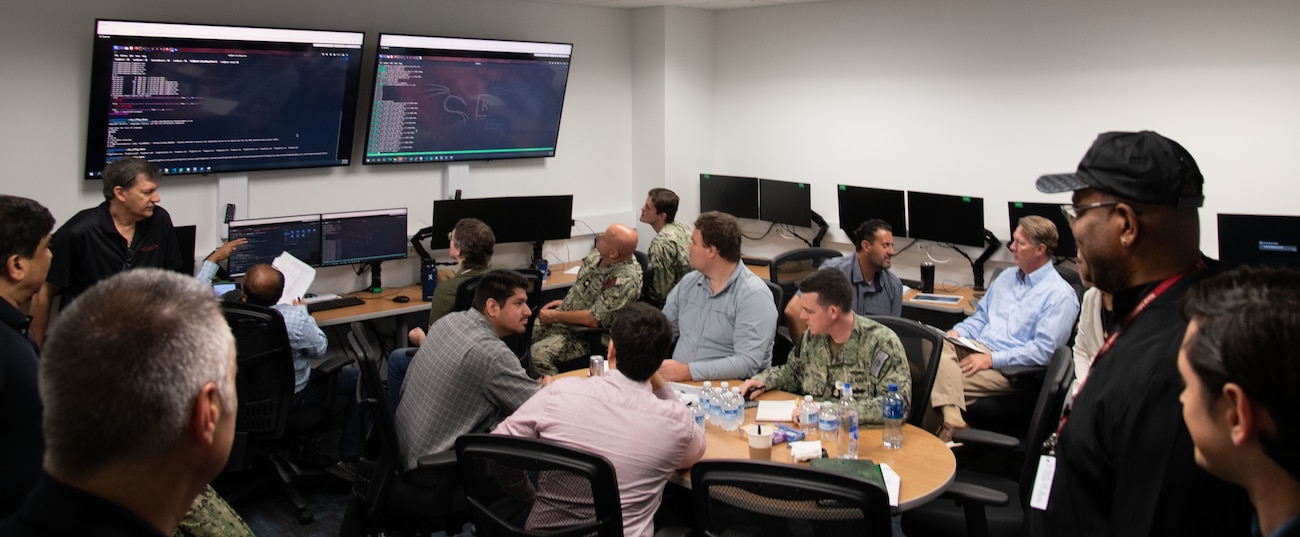 Photo of NIWC Pacific and Atlantic workforce at CyberANTX having a brief discussion.  Woman in black in front of suited men and one male navy seaman in camouflage uniform.