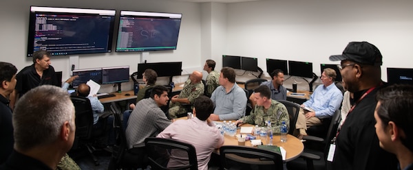 Photo of NIWC Pacific and Atlantic workforce at CyberANTX having a brief discussion.  Woman in black in front of suited men and one male navy seaman in camouflage uniform.
