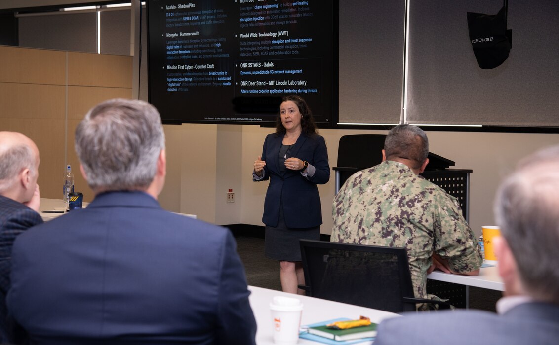 Photo of NIWC Pacific and Atlantic workforce at CyberANTX having a brief discussion.  Woman in black in front of suited men and one male navy seaman in camouflage uniform.