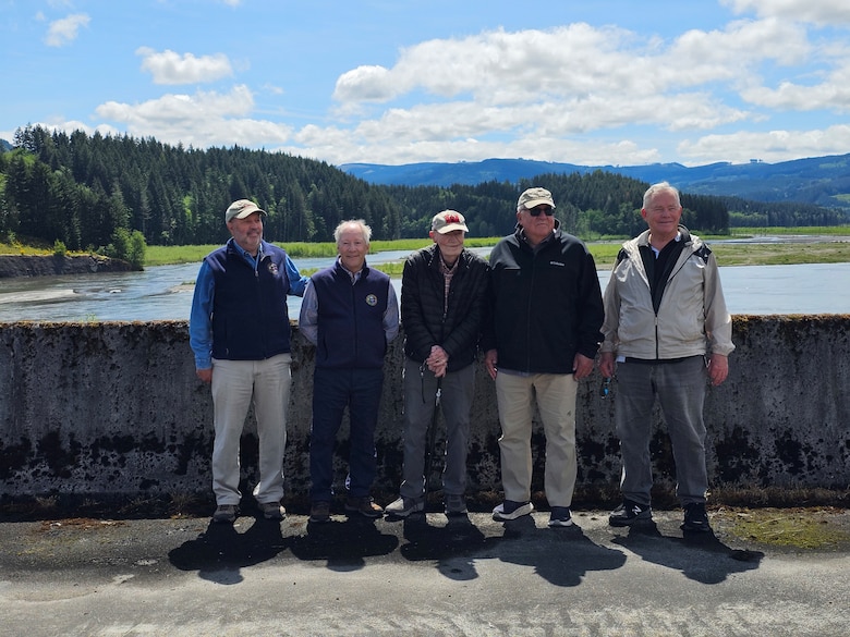 Five men stand on a concrete structure with a river and trees in the background