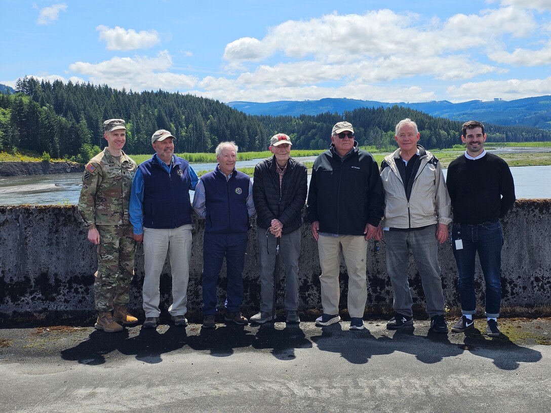 Seven men stand on a concrete structure with a river and trees in the background