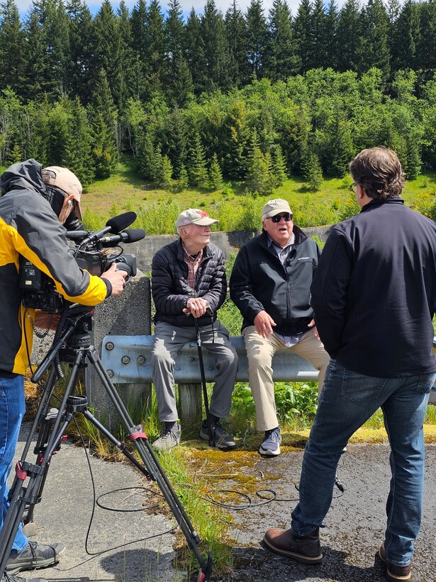 Two men sit on a metal railing speaking to two other men, one with a camera with a forest in the background