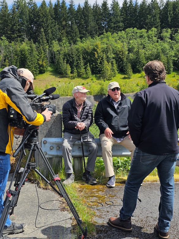 Two men sit on a metal railing speaking to two other men, one with a camera with a forest in the background