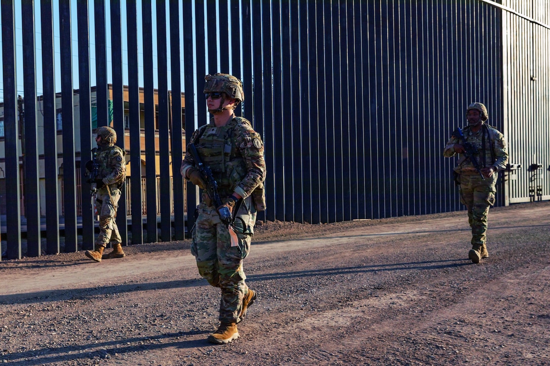 Three soldiers in combat gear walk on a dirt road with a tall security wall on one side; a structure is seen on the other side of the barrier.