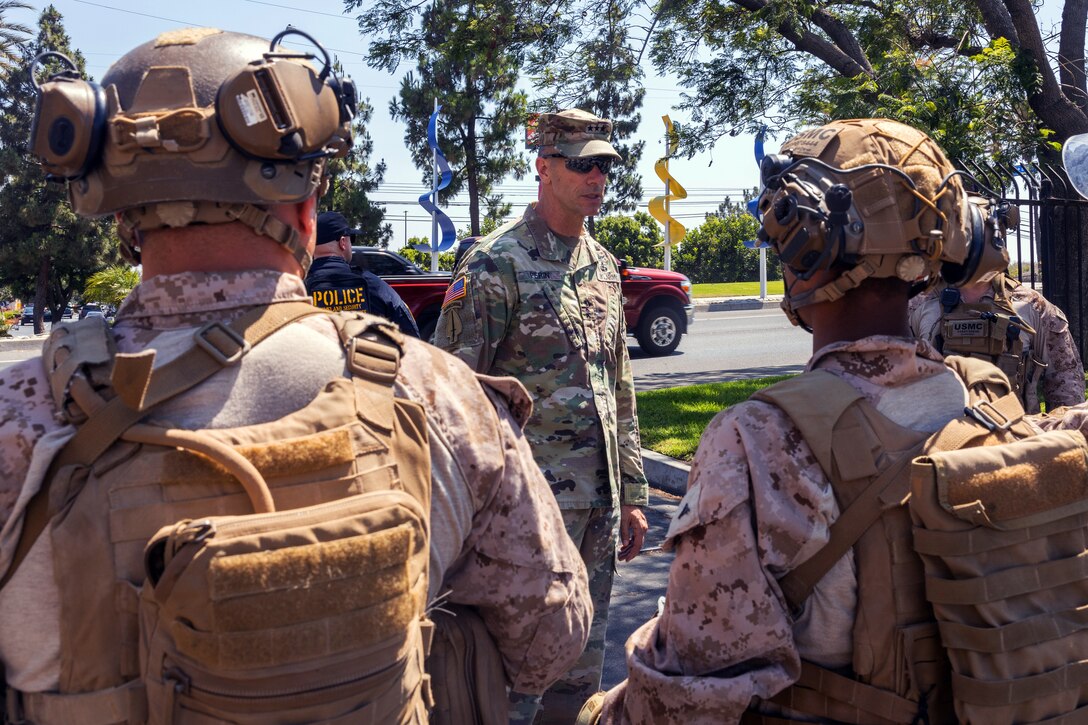 A soldier wearing a cap and sunglasses addresses a group of soldiers in combat gear, with trees and vehicles on a paved lot in the background.