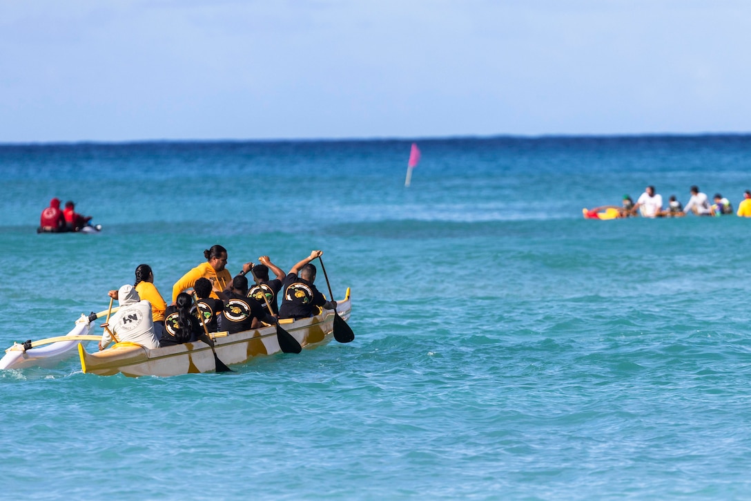 Marines row a boat in calm water on a sunny day, with two people on a Jet Ski-like craft and another group of people seen on a boat in the background.