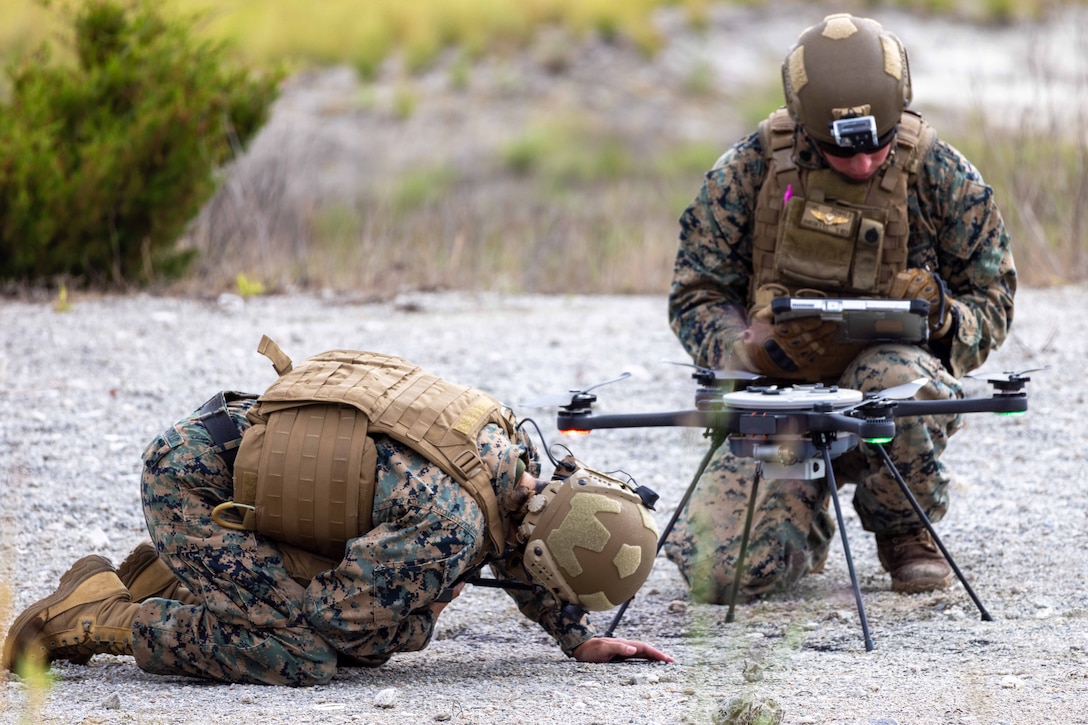 Two Marines, one crouching to look under a drone, inspect the components of the small unmanned aircraft system in a sparsely vegetated area.