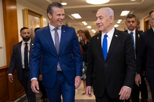 Two men in business suits are smiling as they walk and talk in a large hallway. There are three additional men in business suits walking behind them in the background.